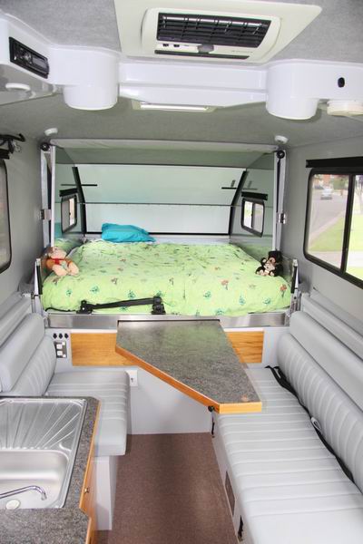 Vanaf de deur. 
De slaapkamer (160x200) en de zit-/eet-/TVkamer (de zwarte steun bij het bed is voor de TV, maar hebben we verwijderd). 
De tafel schuift van onder het bed vandaan en kan op verschillende standen vrijhangend vastgezet worden (hij kan nog verder uitgeschoven worden dan op de foto) 
Onder de korte bank zit bergruimte, onder de lange bank zit deels bergruimte met een buitenluik naast de deur, deels de kachel en deels de BBQ / Wokbrander met buitenluik. Bovenin zit een Radio / MP3 speler met boxen en een airco. 
De airco hebben we in 2013 vervangen door een ventilatieluik, hetzelfde als de andere. 
Tussen de banken slaan we bij een ingeschoven caravan onze 150watt mobiele zonnecellen en onze buitenstoelen en -tafel op, vastgesnoerd.